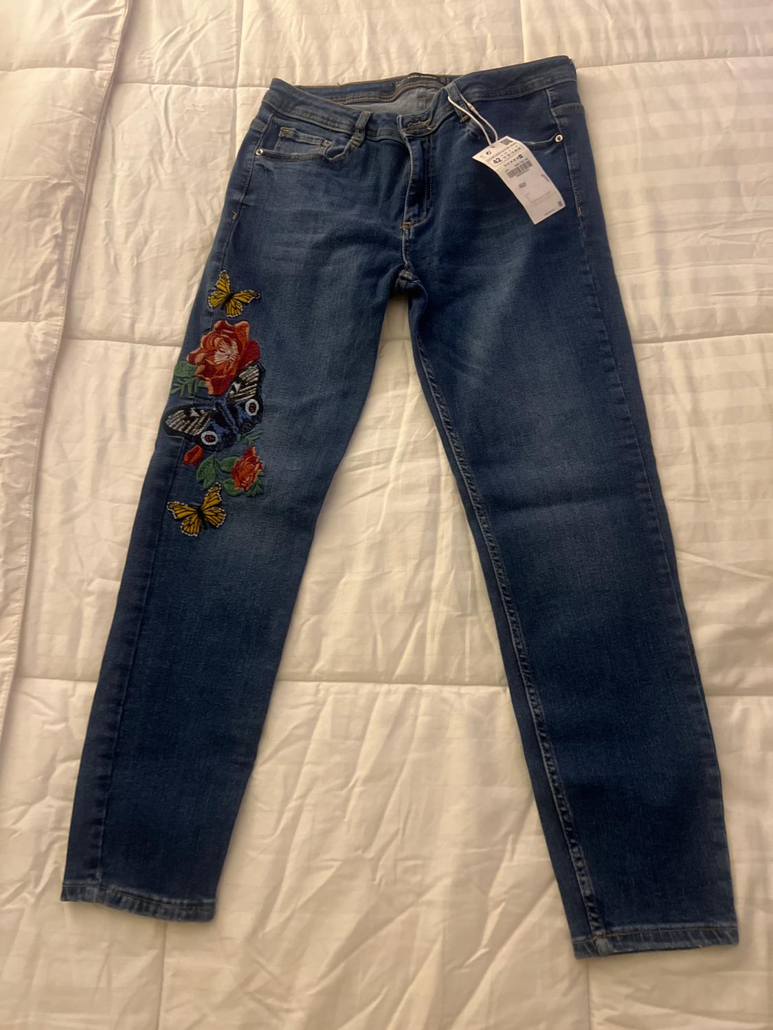 Front view of blue denim jeans with embroidered butterflies and flowers on the left leg.