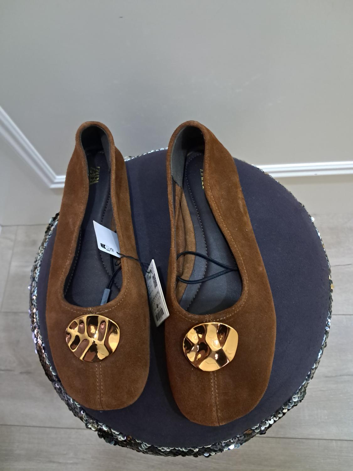 Brown suede flat shoes with gold embellishment on the toe, on a round surface decorated with silver sequins.