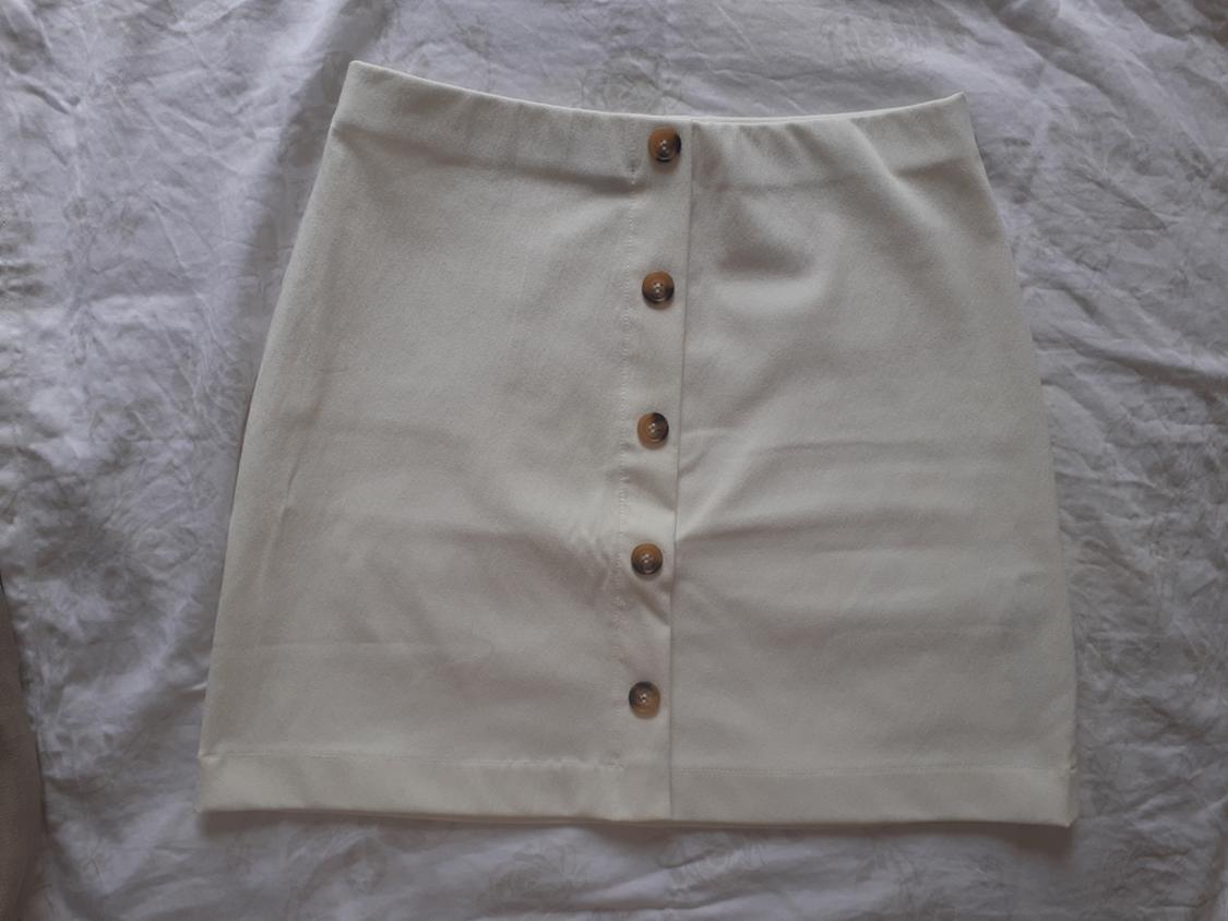 Cream-coloured short skirt with brown front buttons, photographed flat on a crumpled white sheet in a relaxed style.