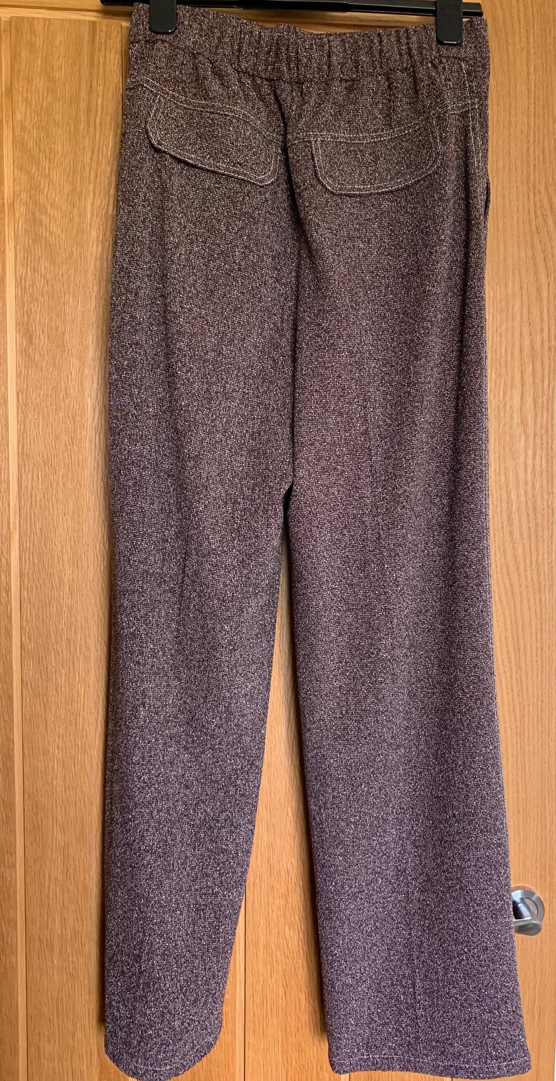 Brown textured fabric trousers hanging on a hanger, with back pockets, against a light wooden background.