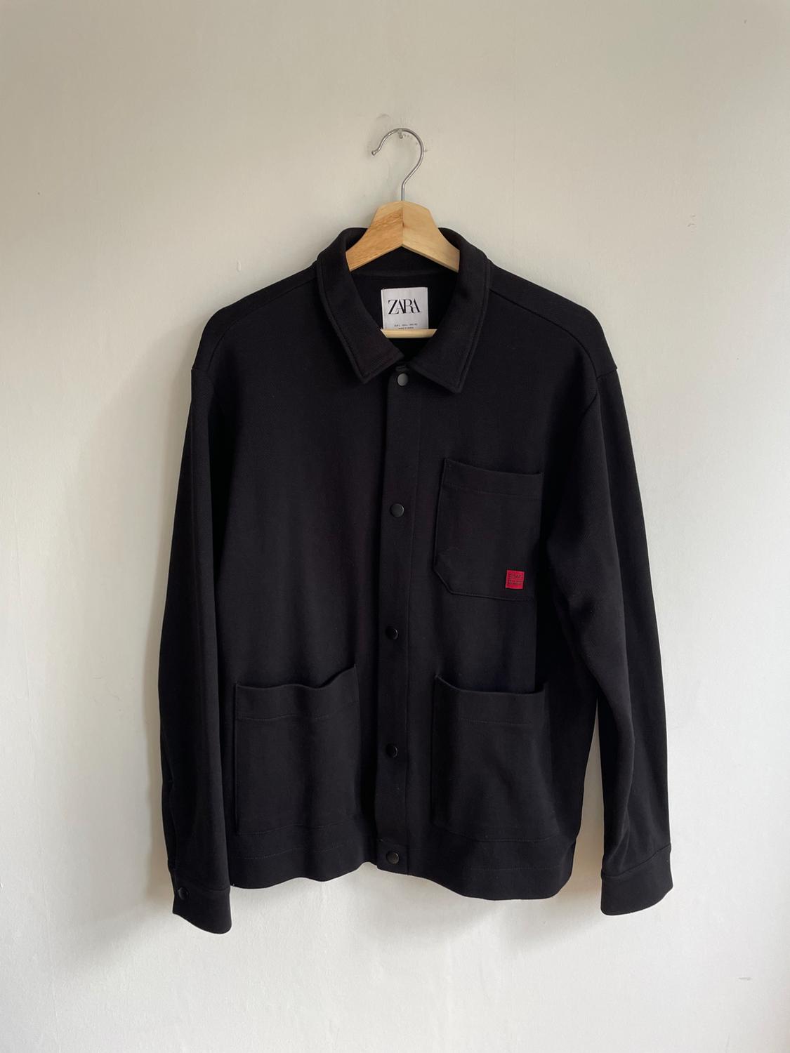 Black jacket hanging on a wooden hanger, with three front pockets and a visible brand label on the collar.