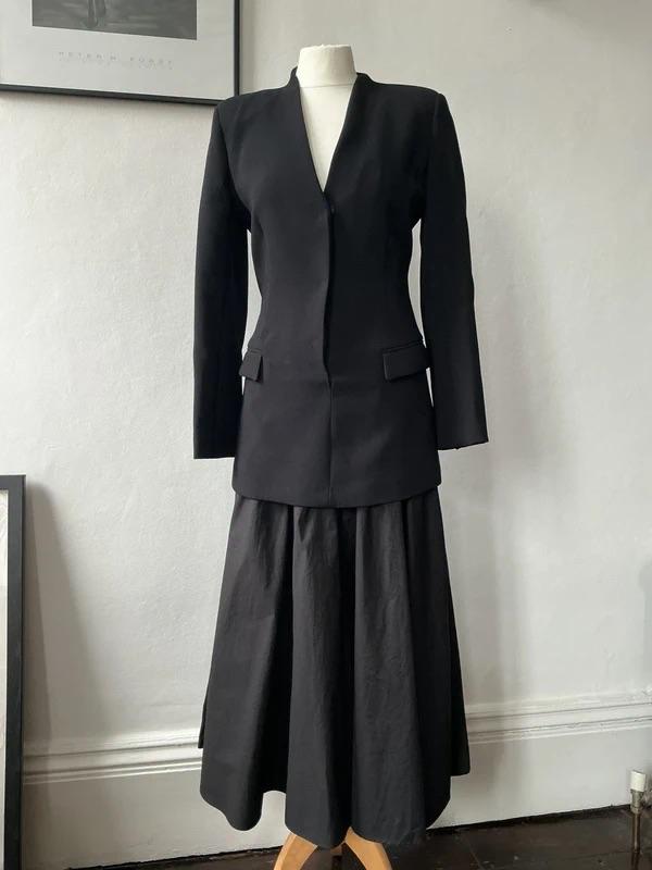 Mannequin wearing a minimalist black outfit with a fitted collarless jacket and a pleated midi skirt, inside a white room.