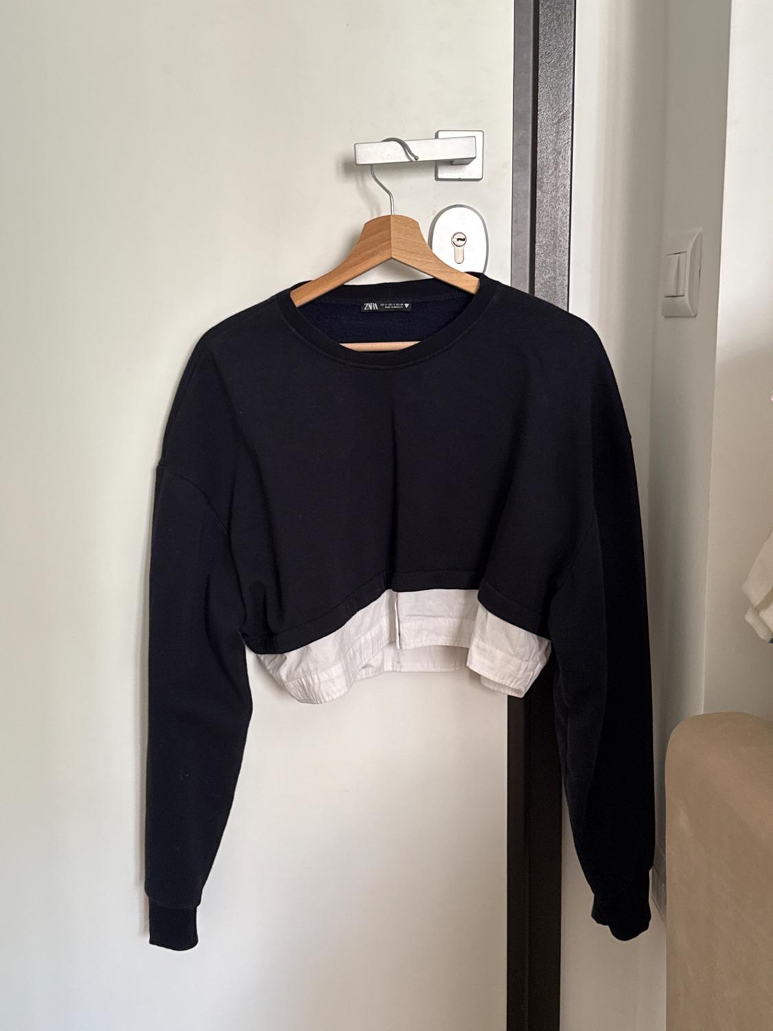 Black, long-sleeved, cropped sweatshirt with a white shirt-style hem, hanging on a wooden hanger on a white door. ZARA label.