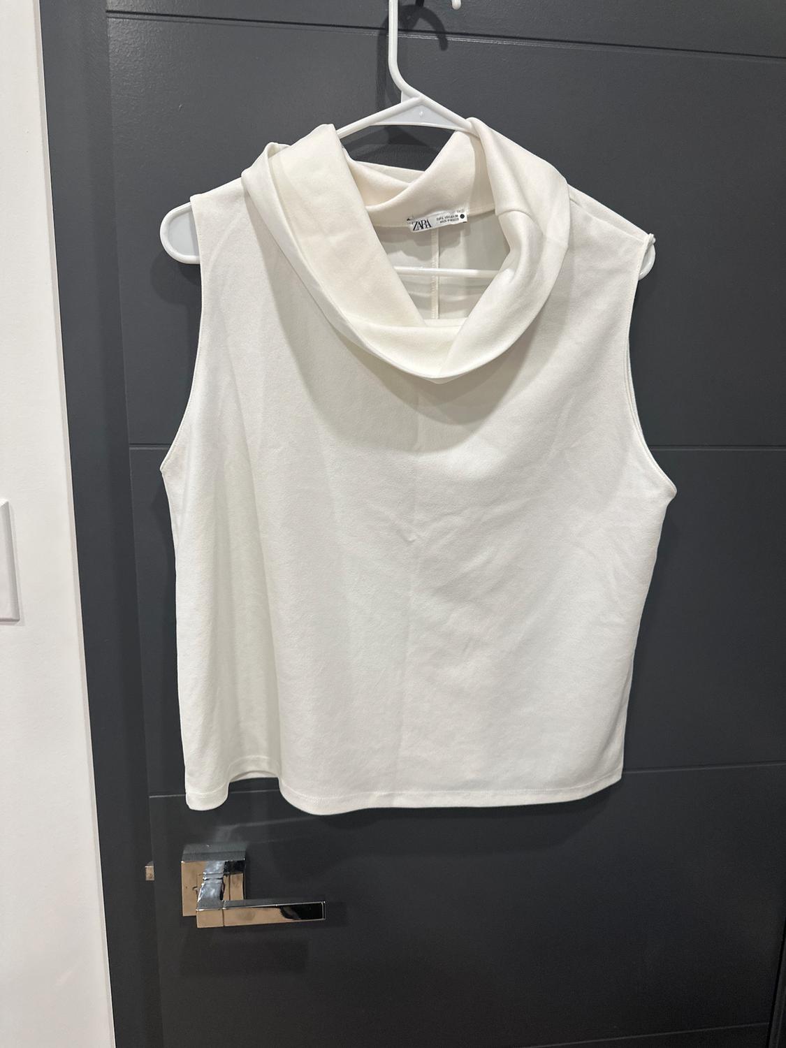 White sleeveless blouse with a high collar hanging on a white hanger over a black door. Tag visible on the inside.