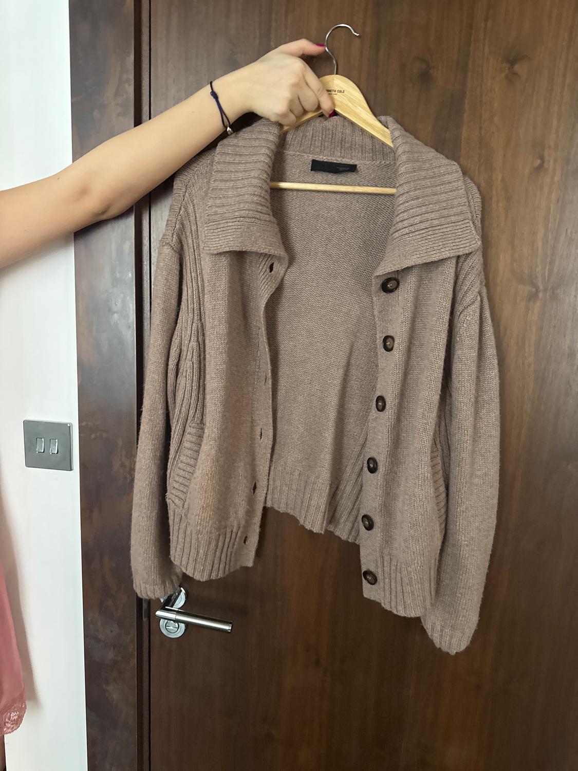 Hand holding a chunky brown knitted cardigan on a wooden hanger, in front of a dark wooden door.