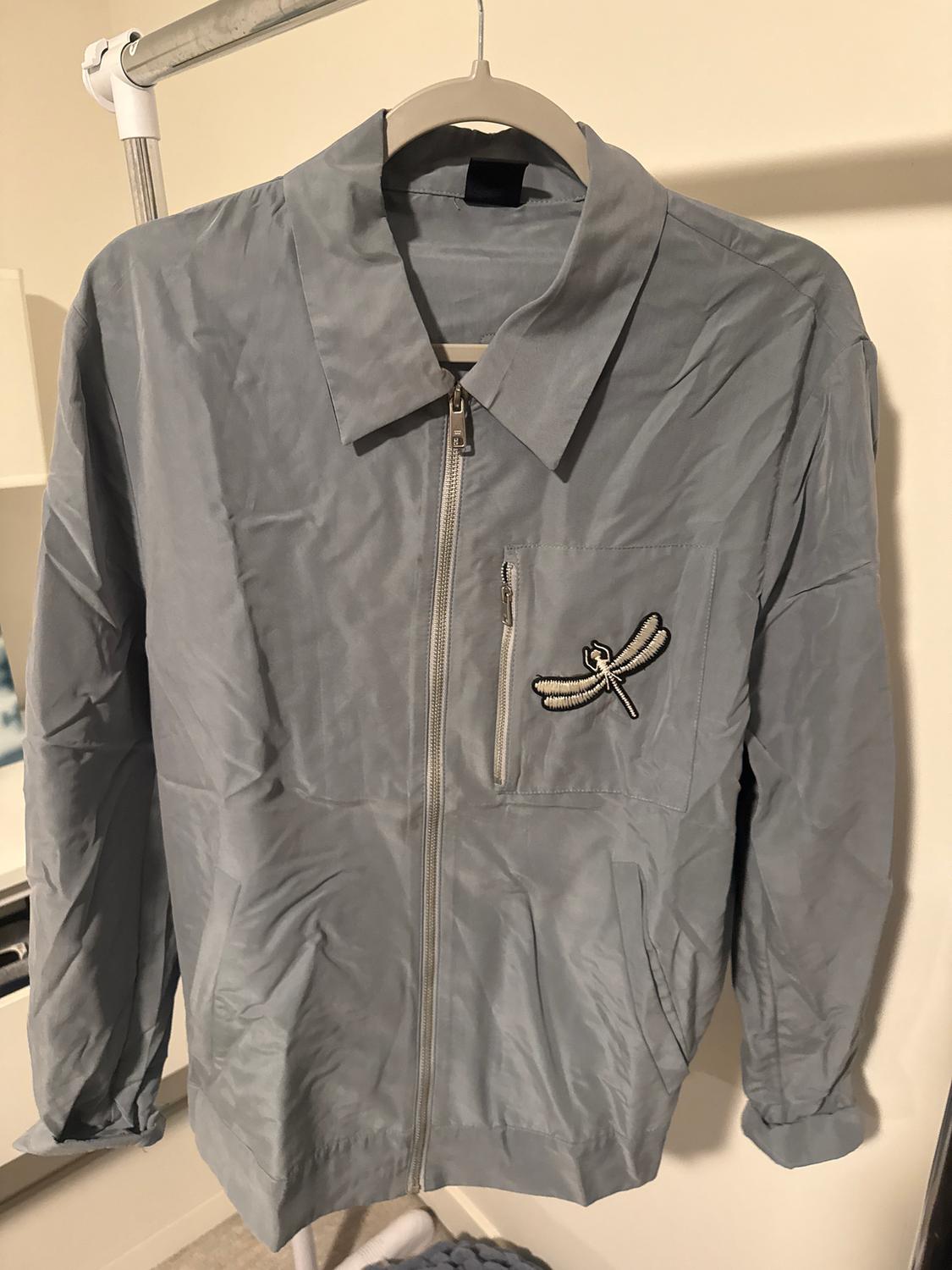 Light grey zip-up jacket with dragonfly embroidery on the chest. Hung on a hanger against a neutral background.