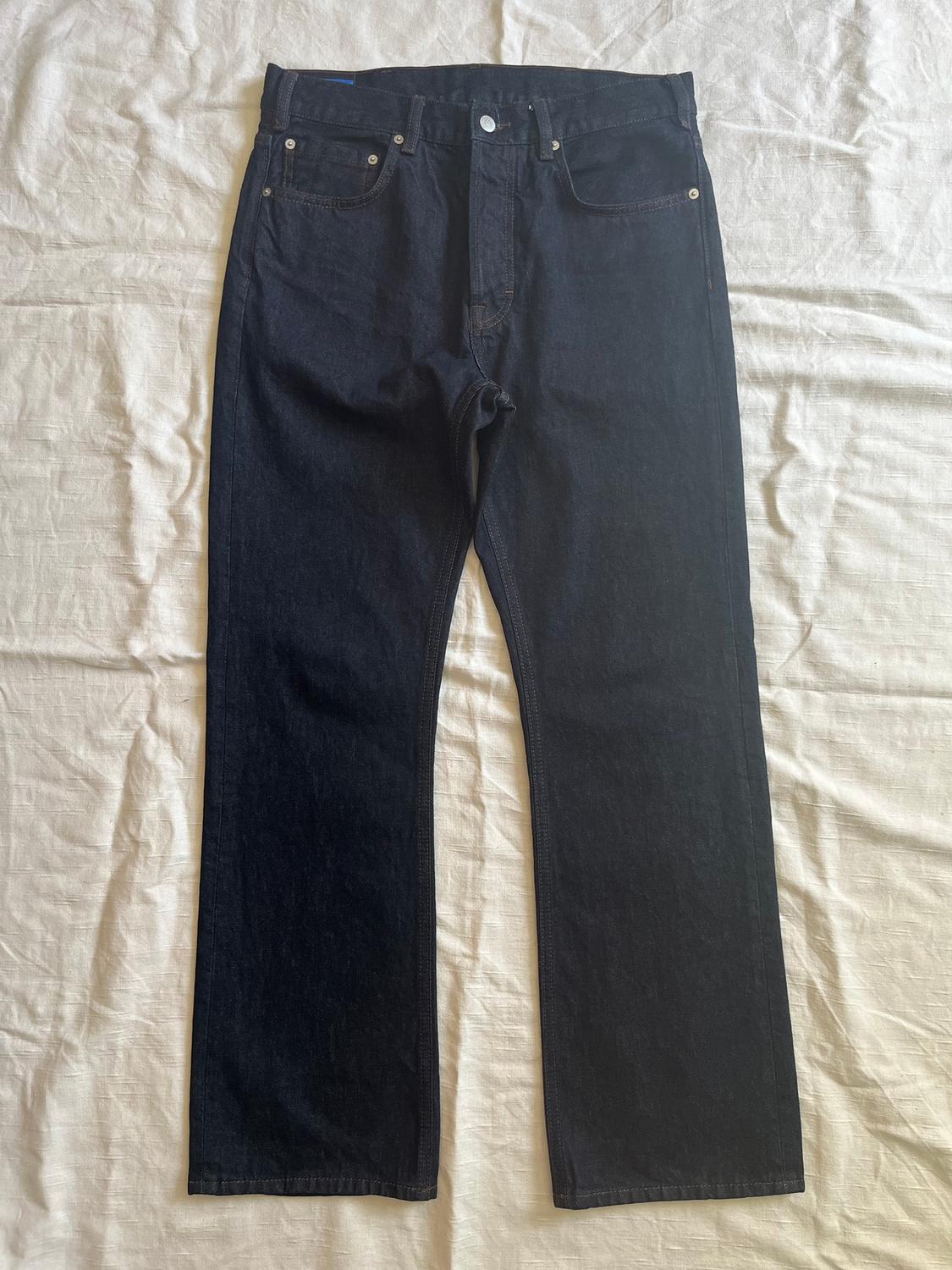 Dark, straight-cut jeans on crinkled white fabric, with five pockets and a metallic button fastening.
