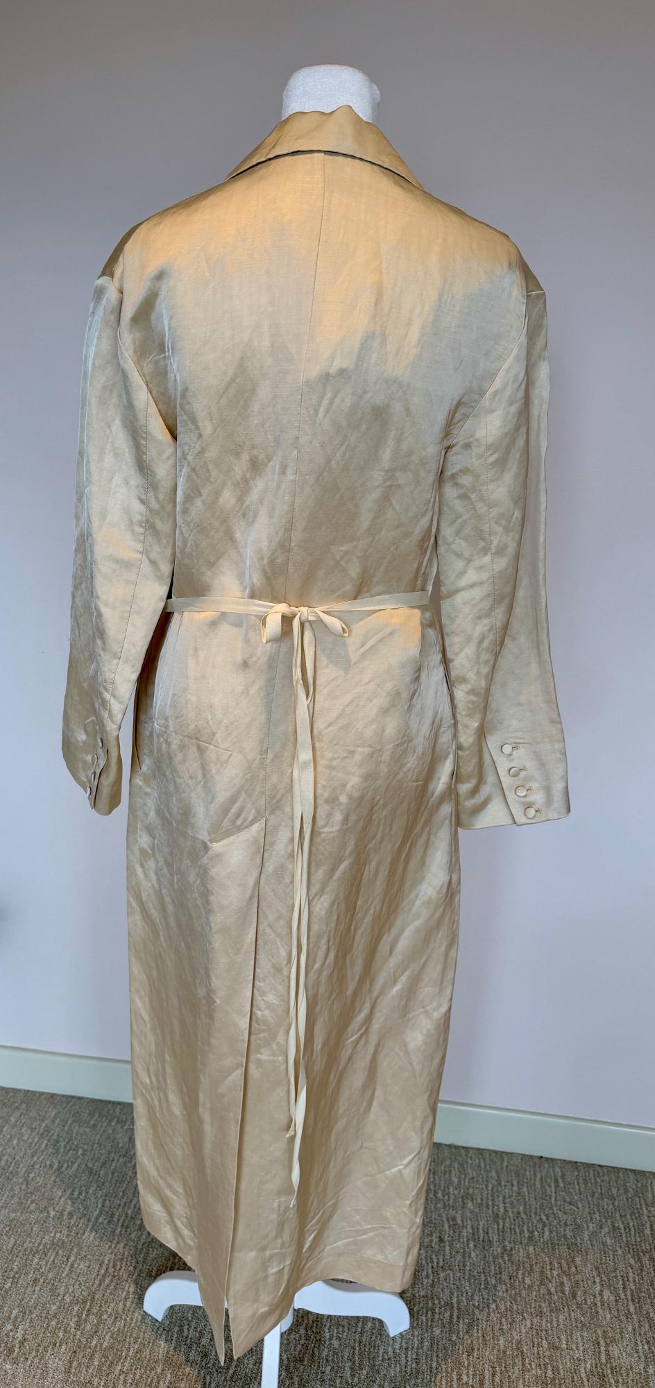 Long beige coat on a mannequin, viewed from the back. It has a thin belt and buttons on the sleeves. Neutral background and carpeted floor.