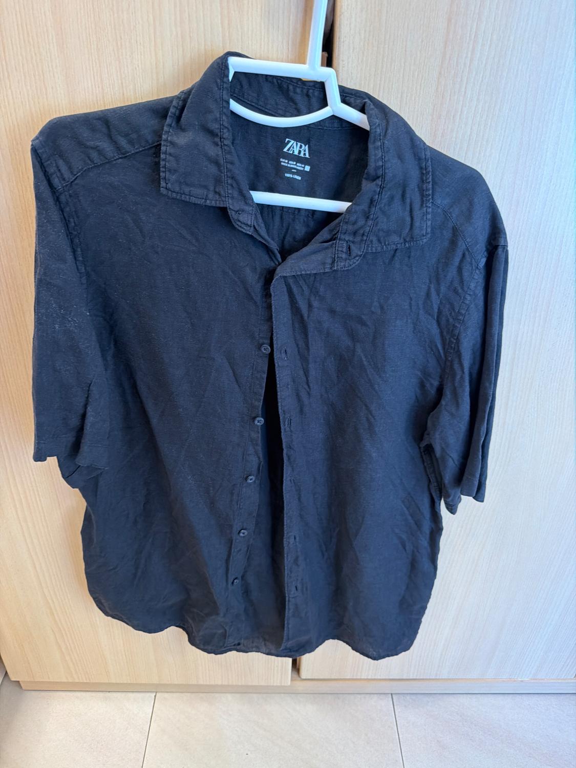 Casual black short-sleeved shirt hanging on a white hanger in front of a light wood wardrobe. Visible label with the text ZARA.