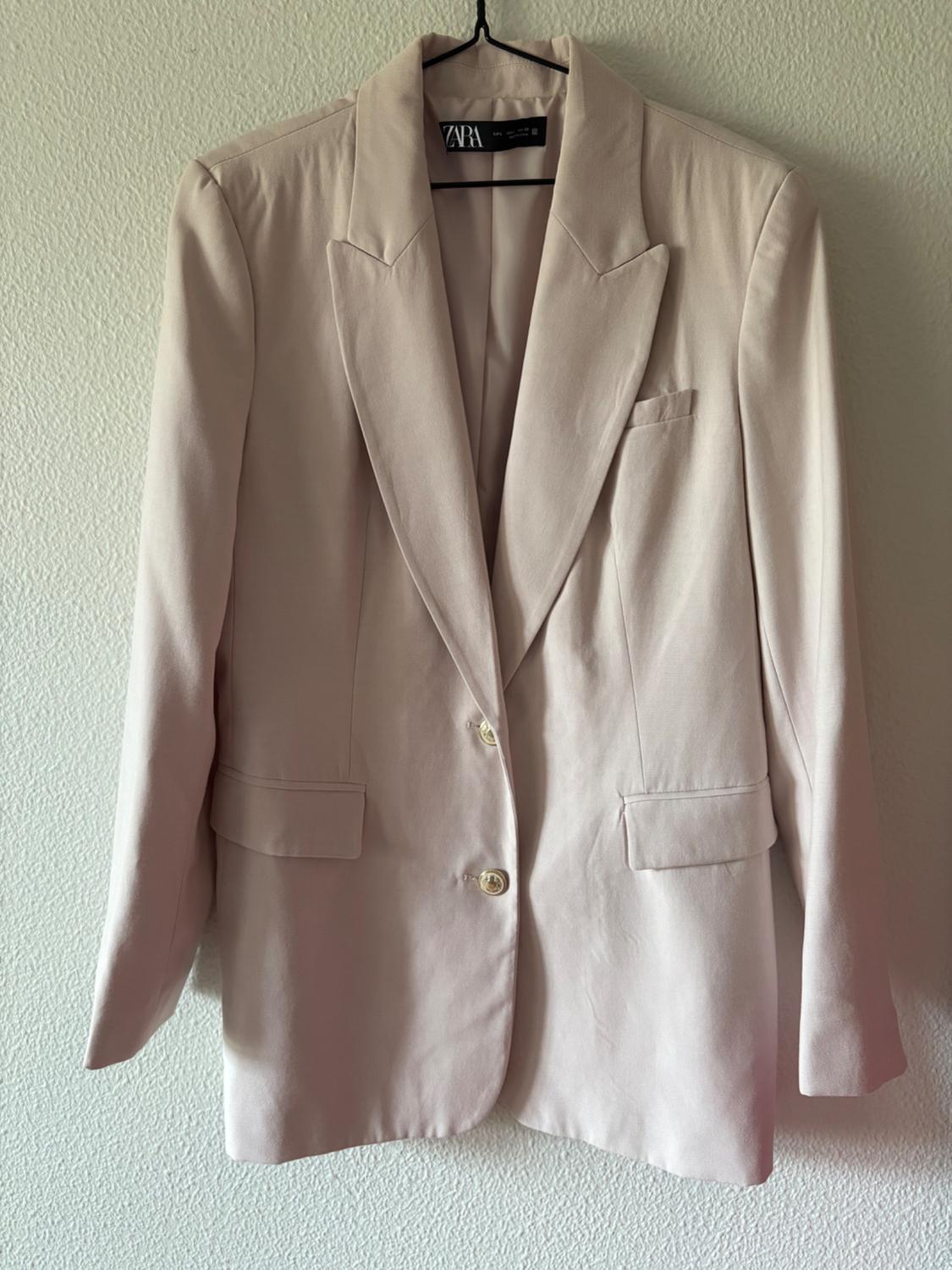 Beige jacket hanging on a hanger, with wide lapels and two front buttons. Visible label with the brand "ZARA".