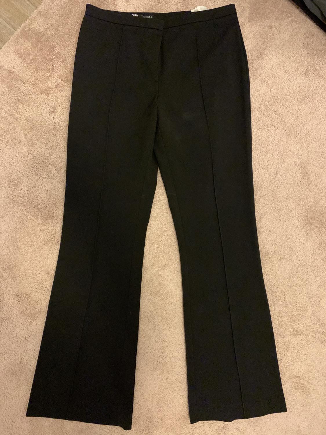 Black dress trousers with front pleats, placed on a beige rug. Label visible at the waistband.