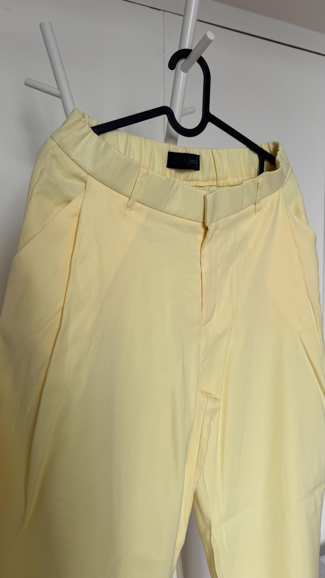 Light yellow trousers hanging on a black hanger on a white clothes rack. Detail of elasticated waist and label with the text ZARA.