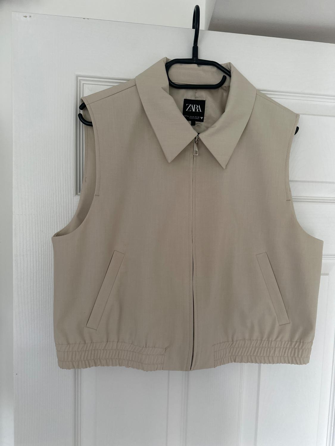 Beige sleeveless gilet with zip and classic collar, hanging on a black coat hanger. Label visible: "ZARA". White door background.