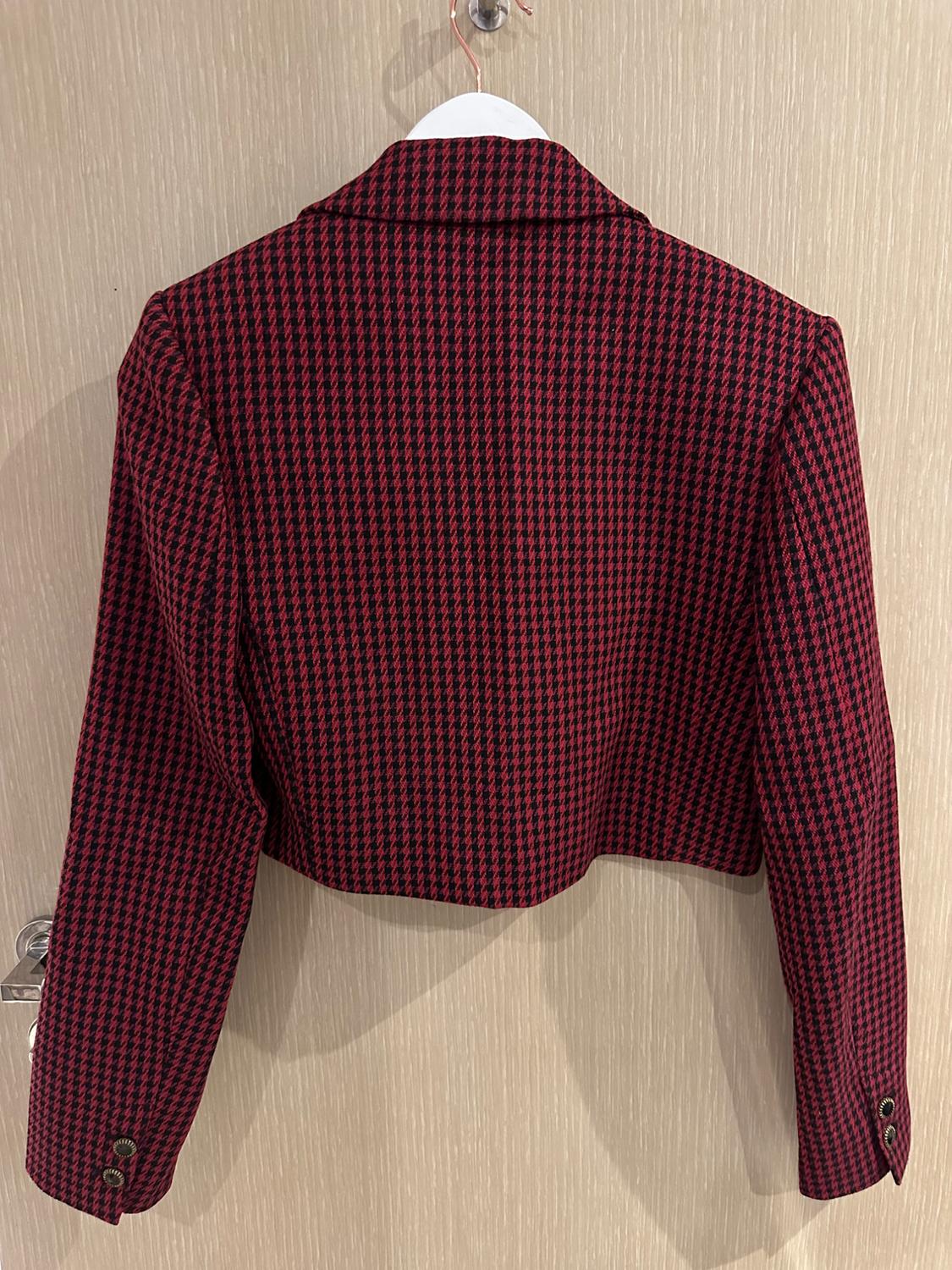 Back view of a short red and black houndstooth jacket, hanging on a white hanger against a wooden door.