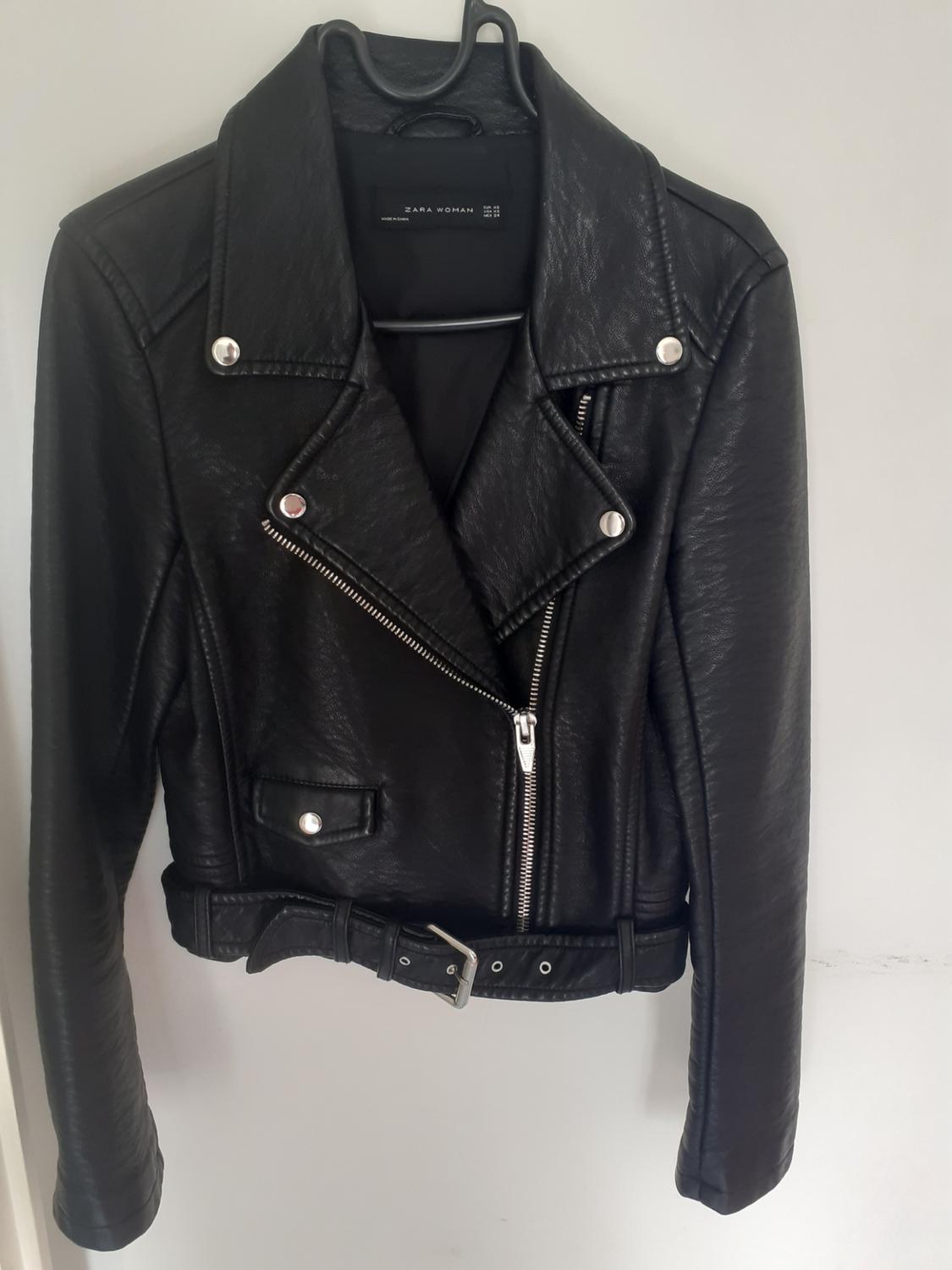Black biker jacket with silver zips and buttons, and a belt at the hem, hanging on a hanger. Visible label: ZARA WOMAN.