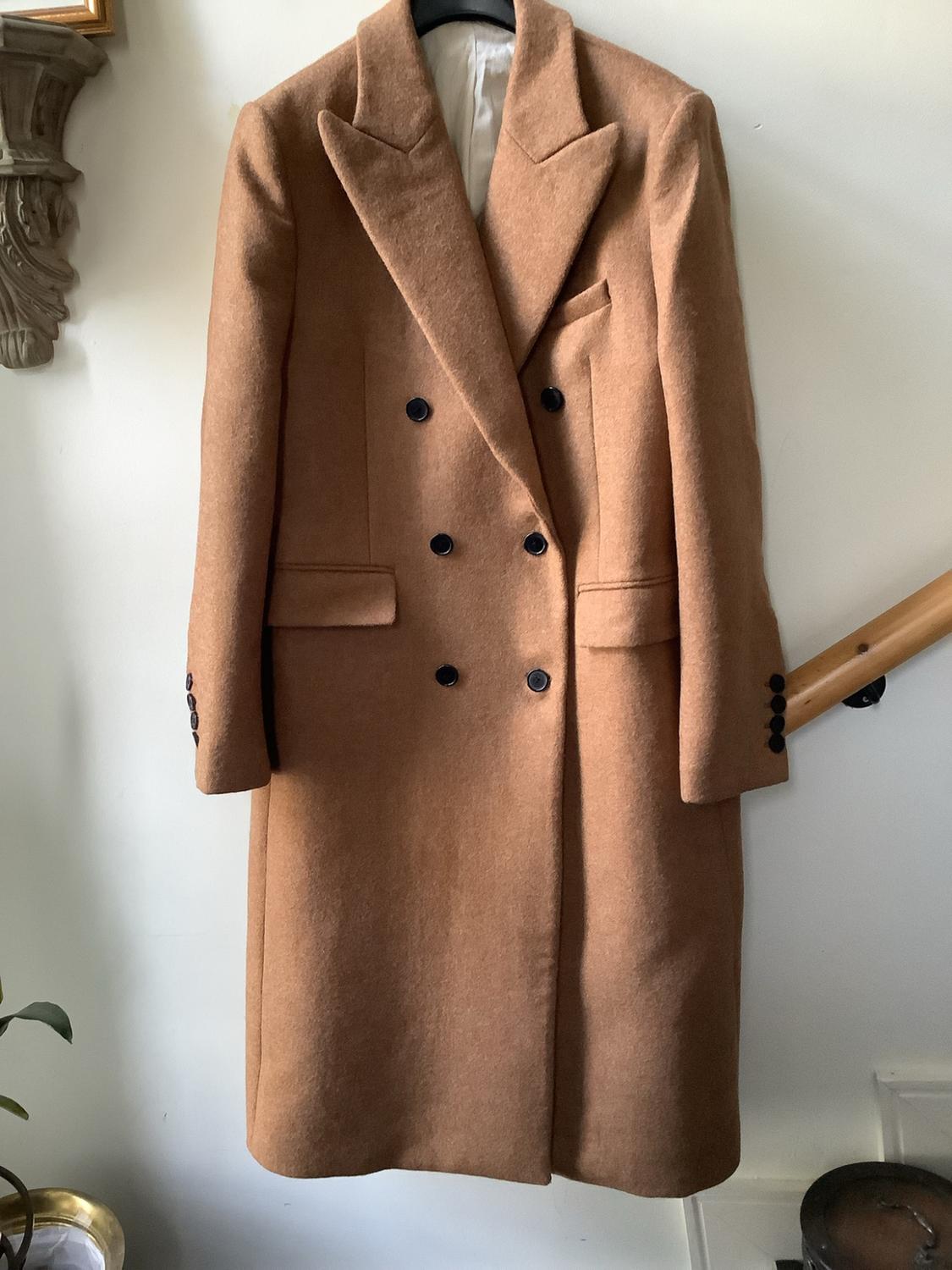 Manteau en laine de couleur camel, à double boutonnage et boutons noirs, suspendu sur un mur blanc.