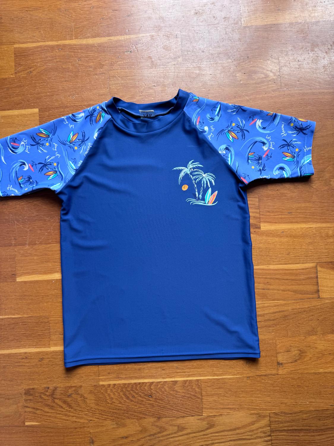 Blue T-shirt with sleeves printed with palm trees and surfboards on a wooden background. Text on sleeves: "Hawaii".