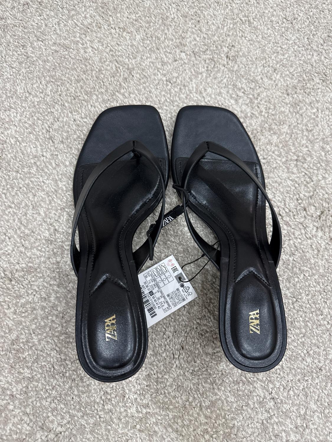 Black heeled sandals with a toe strap on a light-coloured carpet. They have a label and the text ZARA visible on the insoles.