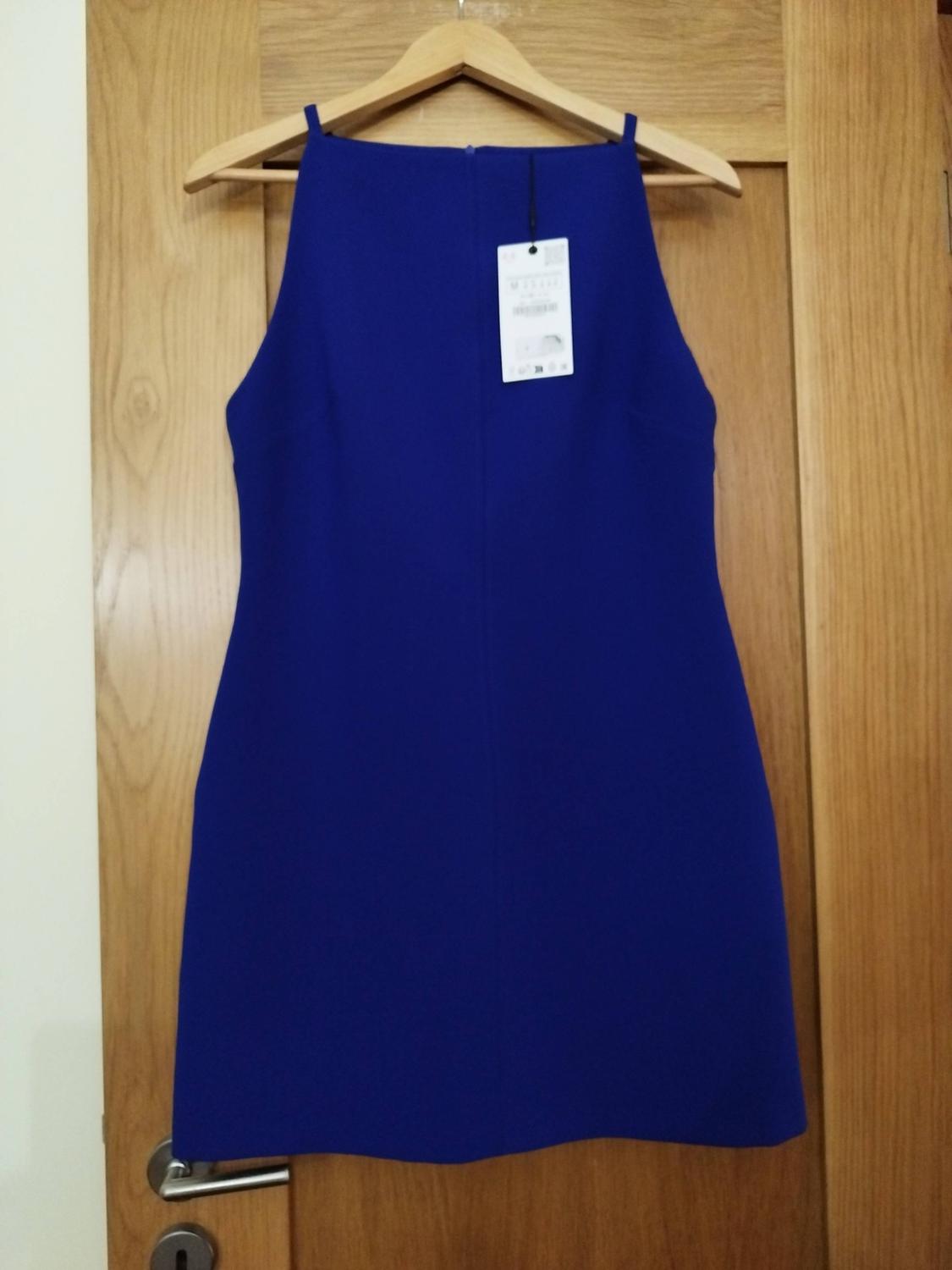 Intense blue short dress with thin straps and a tag, hanging on a hanger over a wooden door.