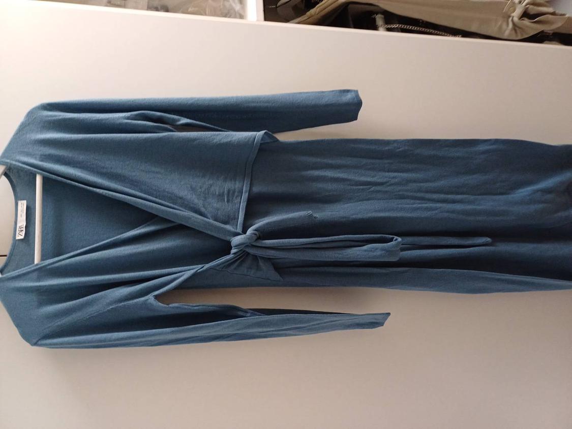 Blue long-sleeved dress hanging on a white hanger against a light surface. It has a belt tied at the front.
