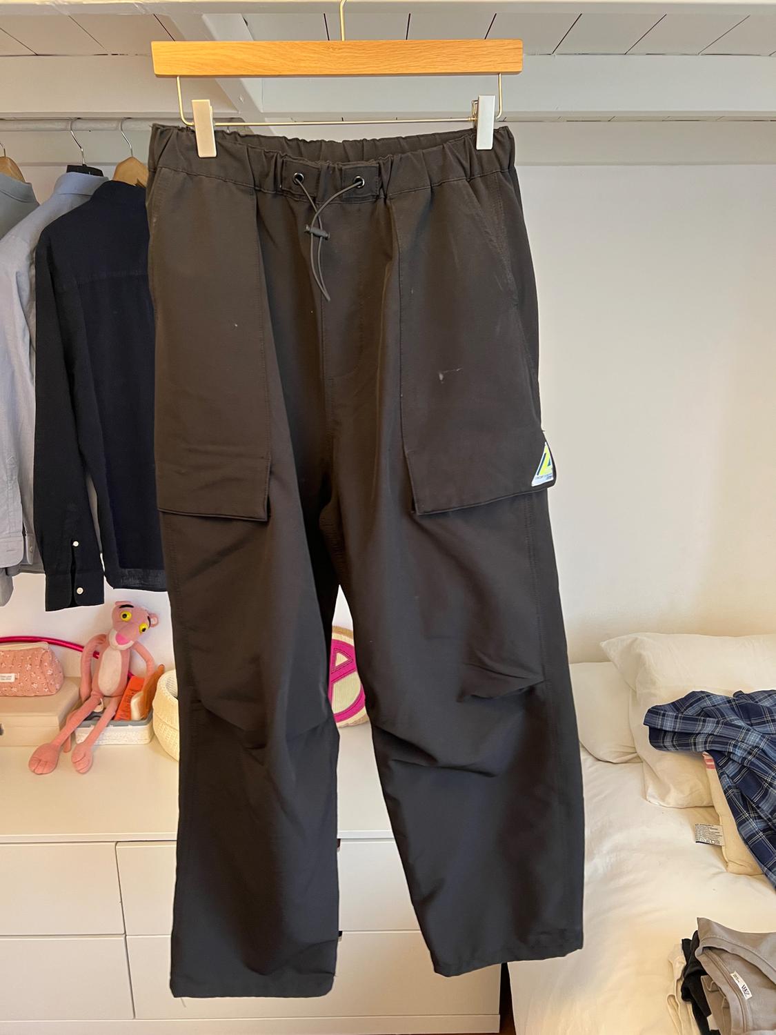 Black trousers hanging on a wooden hanger in a bedroom. Background with shirts hanging and a messy bed.