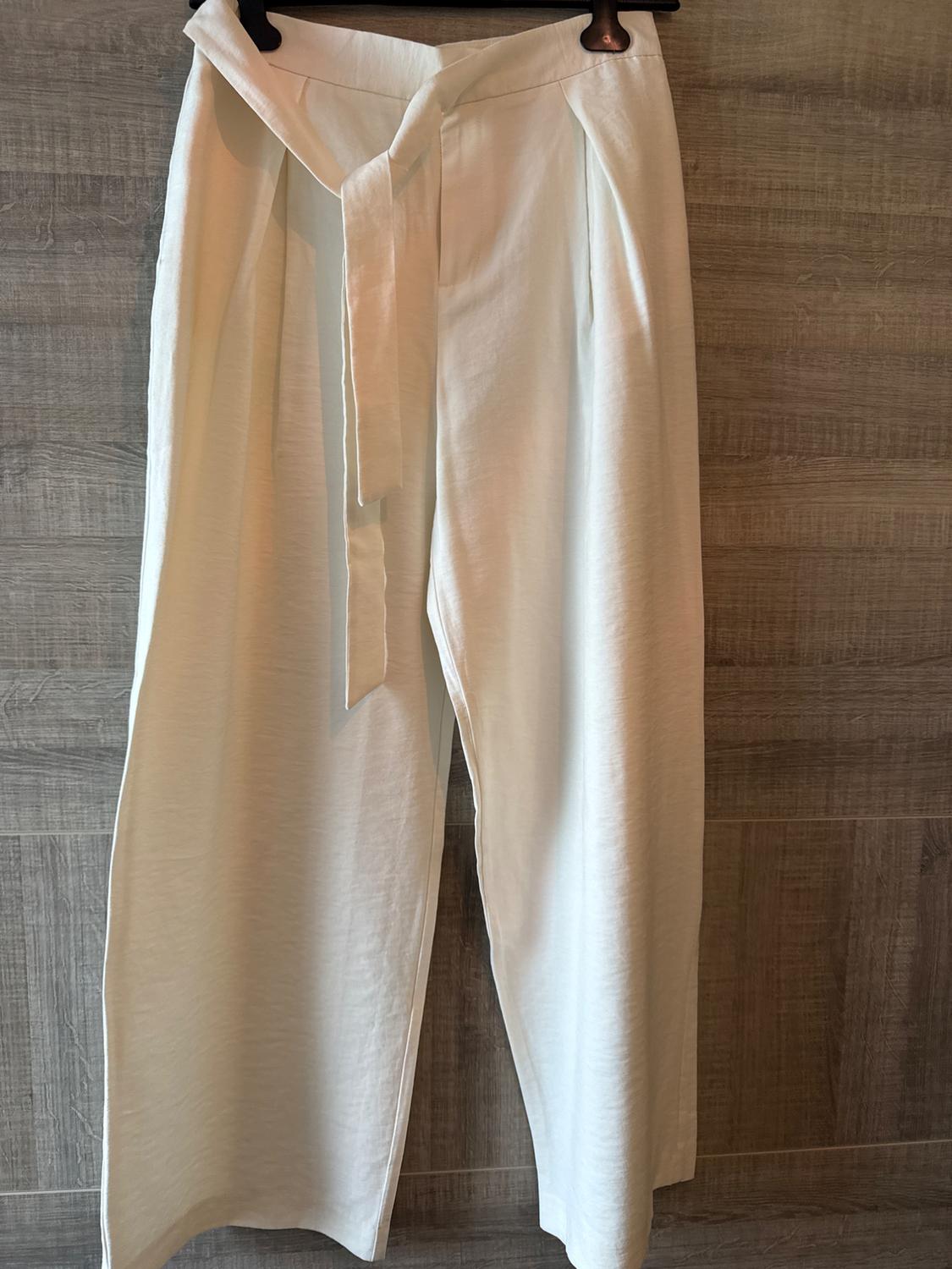 White lightweight fabric trousers hanging on a hanger, with a belt tied at the front, against a grey wooden background.