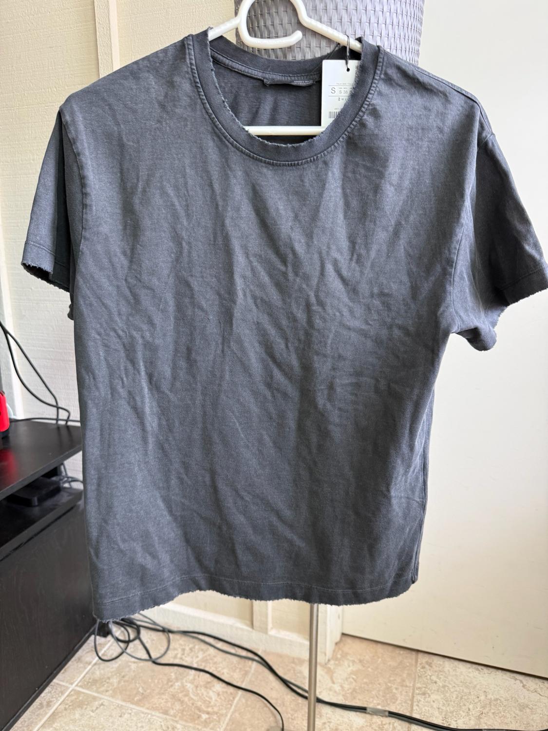Dark grey short-sleeved T-shirt with frayed edges, hanging on a white hanger in a room with cables on the floor.