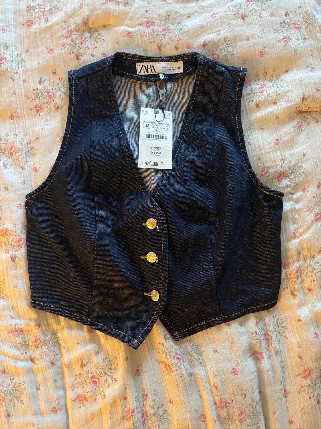 Dark blue denim waistcoat with gold buttons on a floral quilt. Visible label with price and size from Zara.