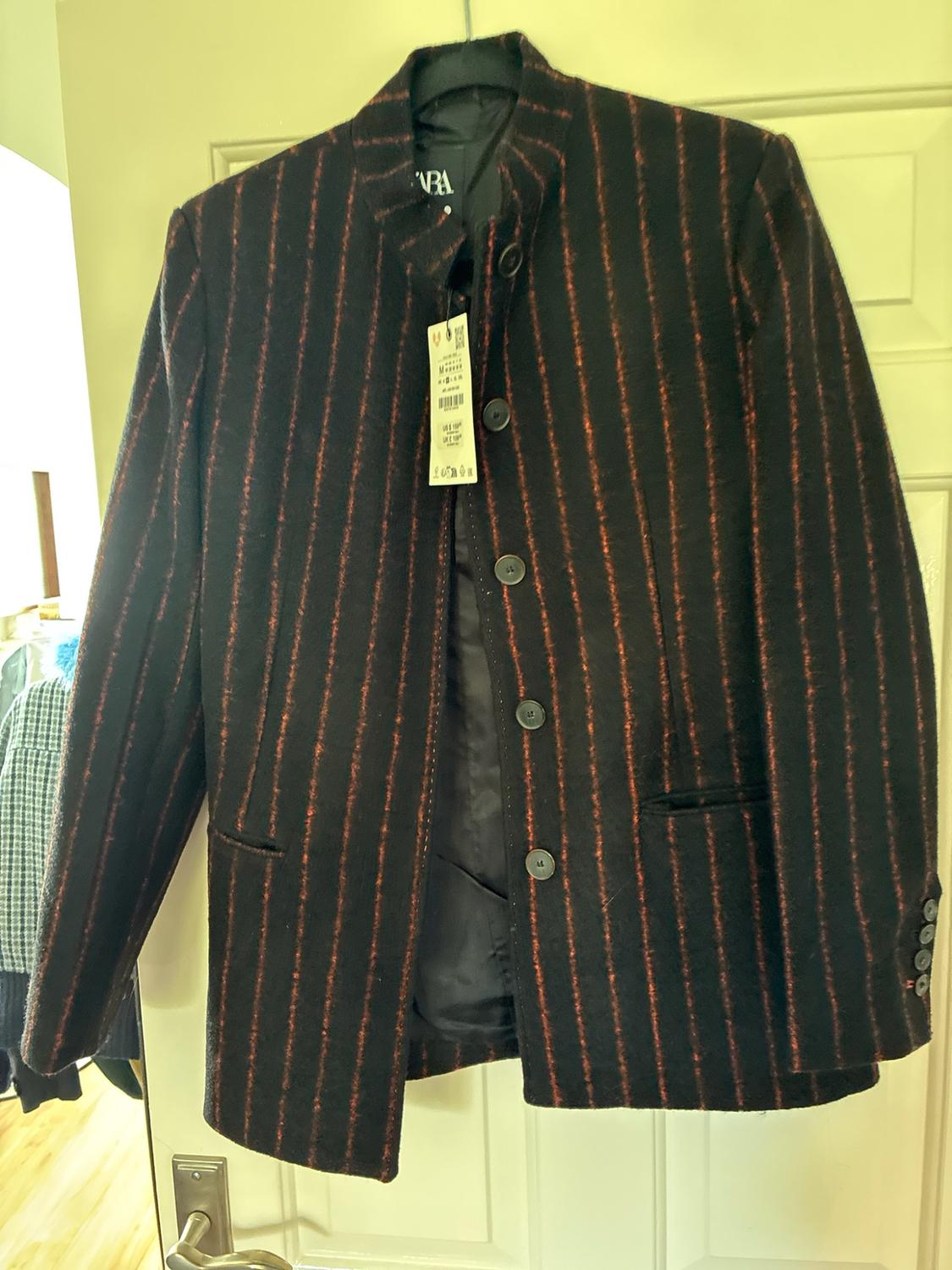 Black jacket with red vertical stripes, front buttons and a label, hanging on a hanger over a white door.