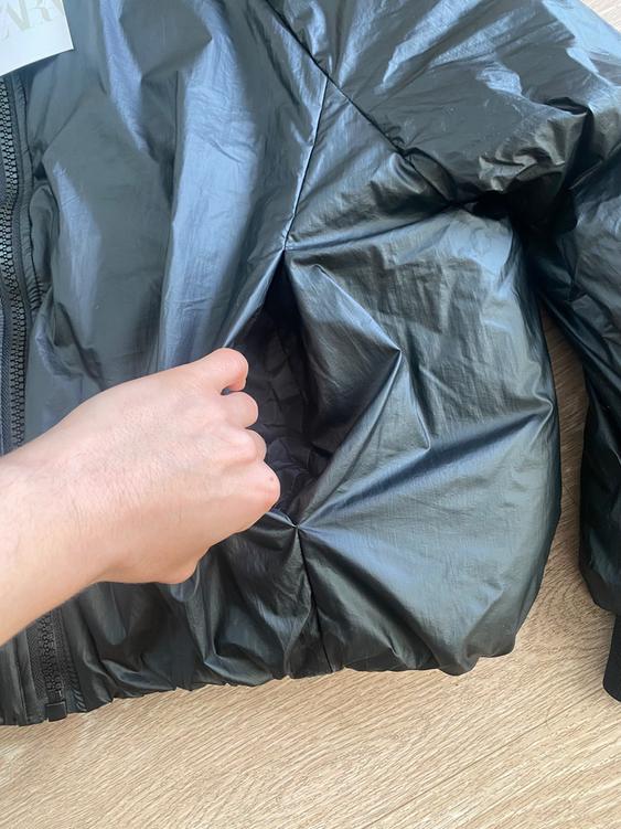 Hand reaching into the pocket of a shiny black jacket on a light wooden surface. Part of a tag visible.