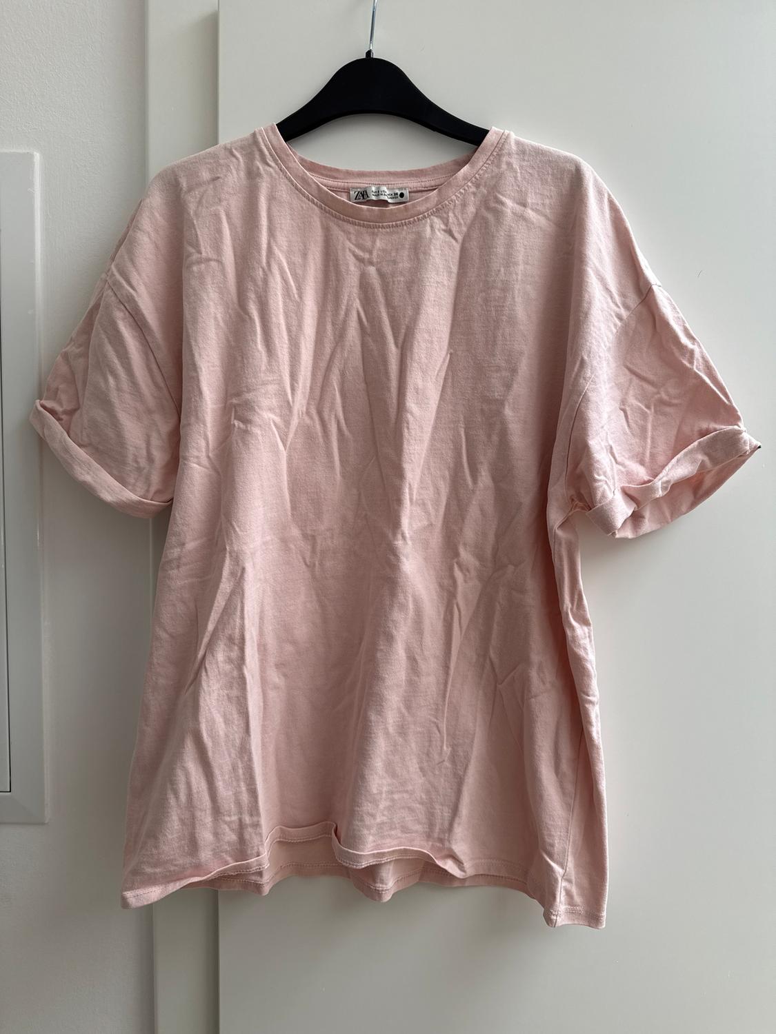 Light pink crumpled T-shirt hanging on a black hanger over a white door. Label visible on the collar.