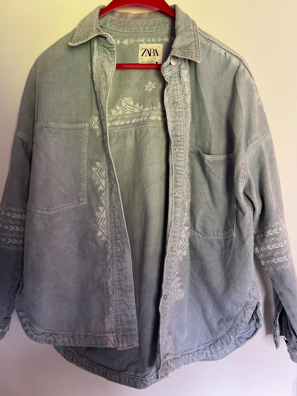 Light blue denim shirt with pockets and white geometric embroidery, on a red hanger against a white background. Visible text: ZARA.