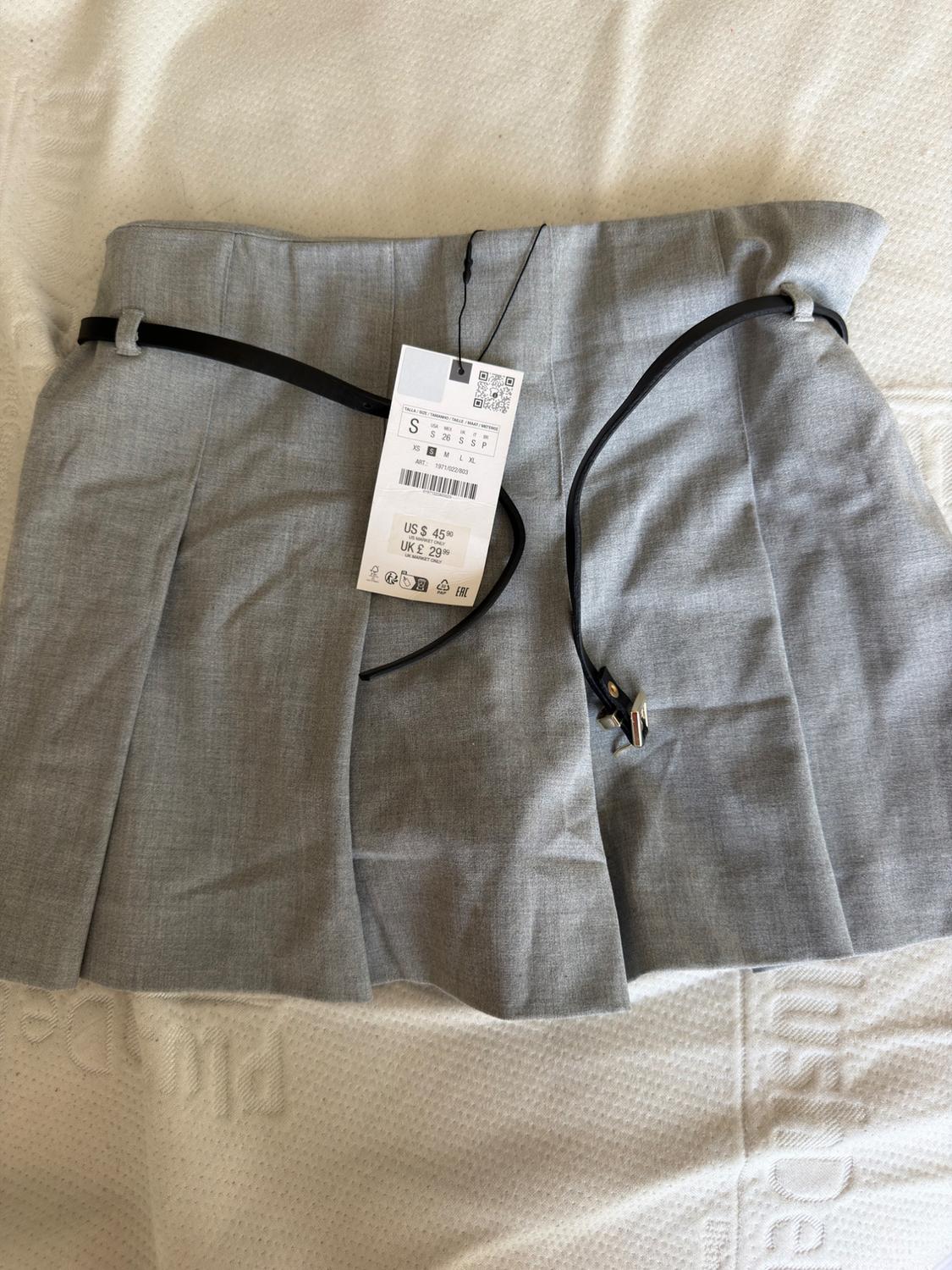 Grey pleated mini skirt with black belt on white surface. Label with text: S US $ 45.90 UK £ 29.99.