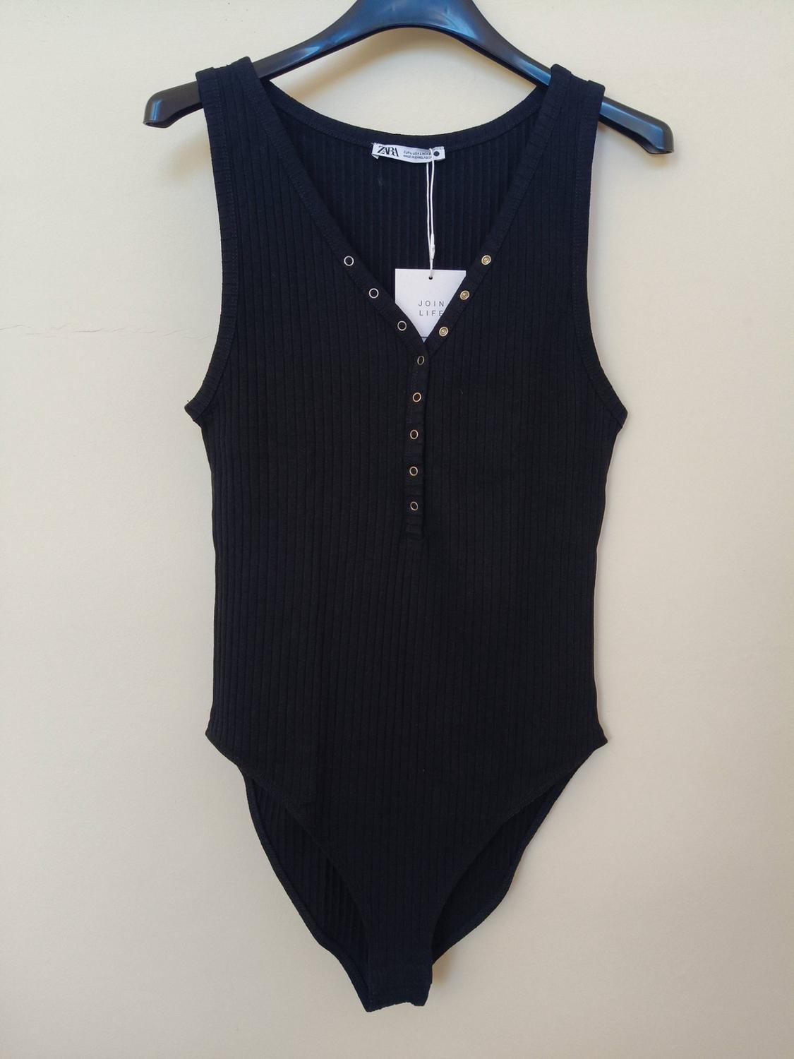 Sleeveless ribbed black bodysuit with button fastening at the neckline, on a hanger against a light background. Labels featuring ZARA and JOIN LIFE text.