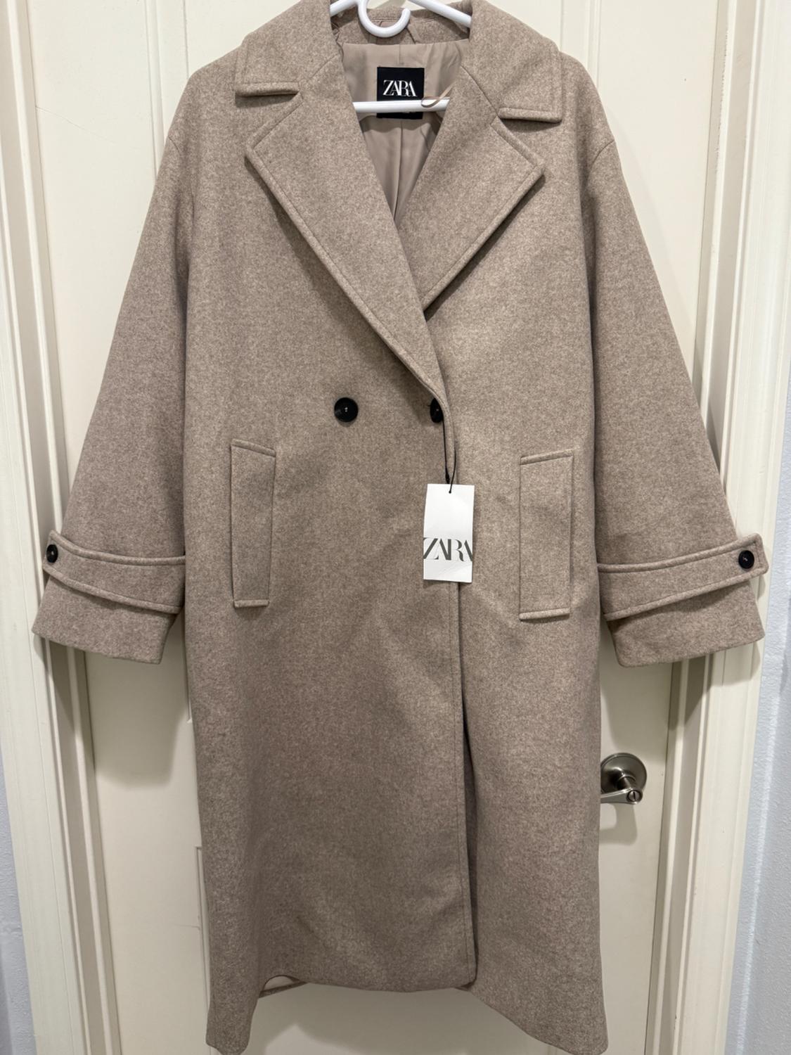 Long beige coat with wide lapels and black buttons, hanging on a white hanger. Visible label with the text "ZARA".