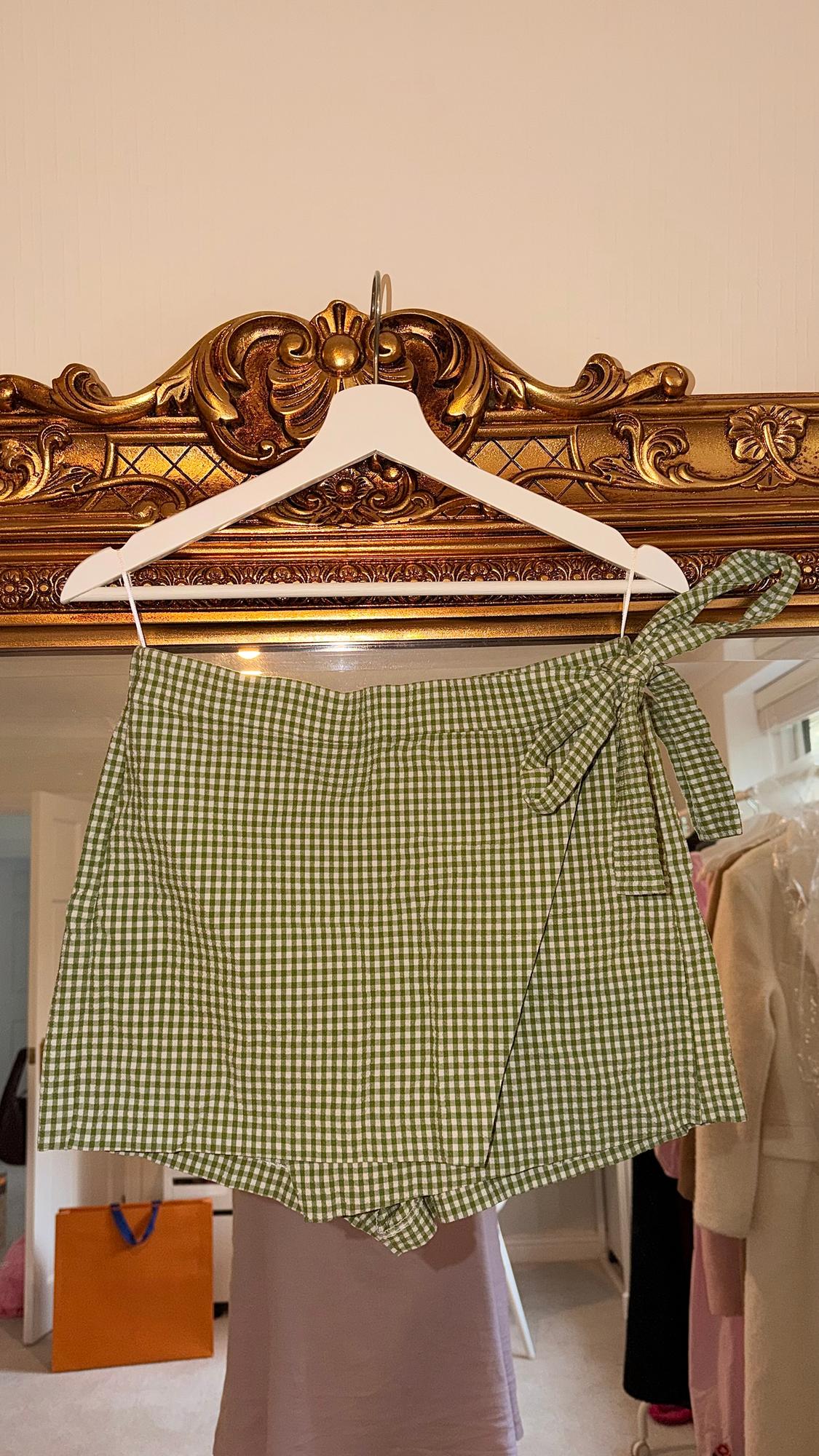 Green and white checked shorts with a bow, hanging on a white hanger in front of an ornate gold-framed mirror.