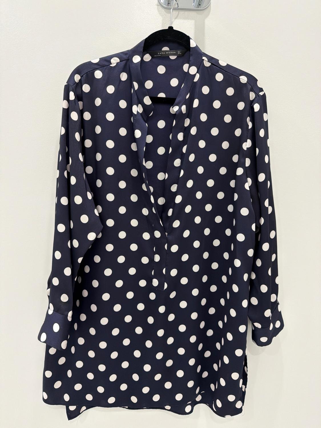 Navy blue long-sleeve blouse with white polka dots, hanging on a black hanger against a white background. Text: ZARA WOMAN.