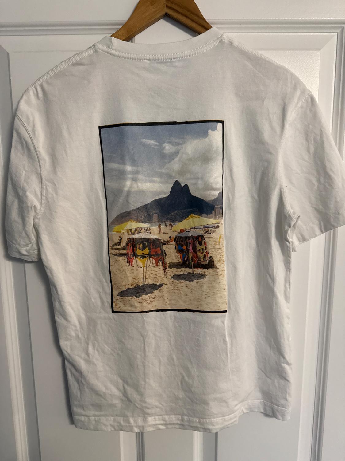 White T-shirt with a beach and mountain landscape print on the back, hanging on a wooden hanger.