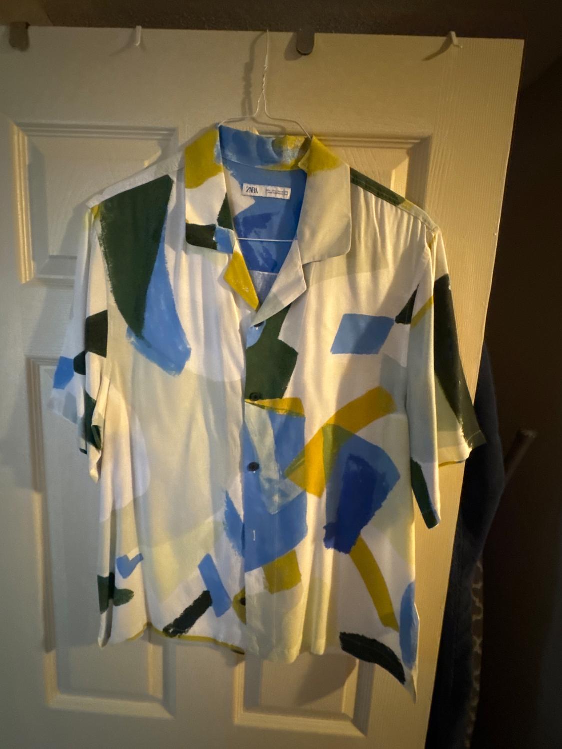 Short-sleeved shirt with an abstract print in shades of blue, green, and yellow, hanging on a white door. Label with ZARA text.