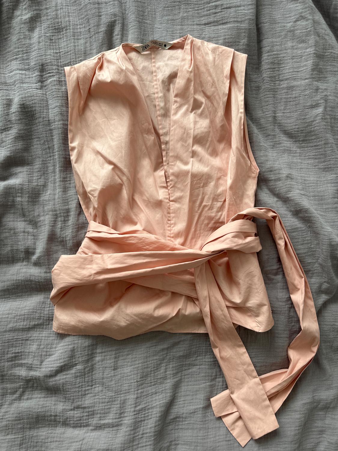 Pale pink, V-neck, sleeveless blouse with a belt, on a crumpled grey fabric. Visible tag: Zara.