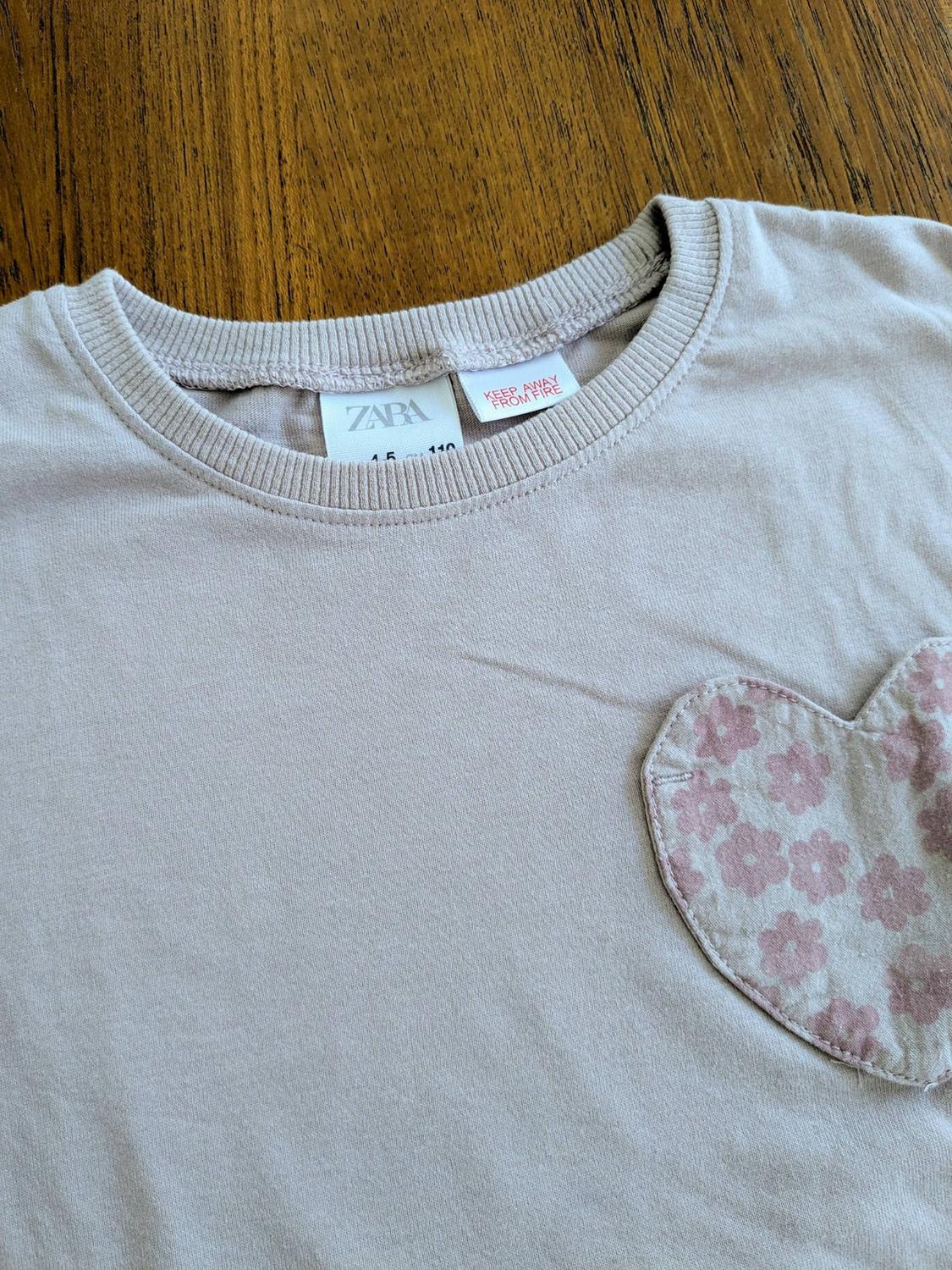 Beige t-shirt with a heart-shaped patch with pink flowers. Visible label: "ZARA" and "KEEP AWAY FROM FIRE". Wooden background.
