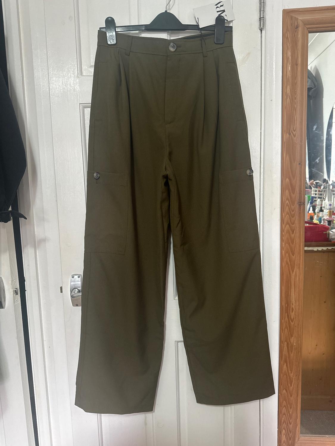 Straight-cut green trousers hanging on a black hanger over a white door. Side pockets visible.
