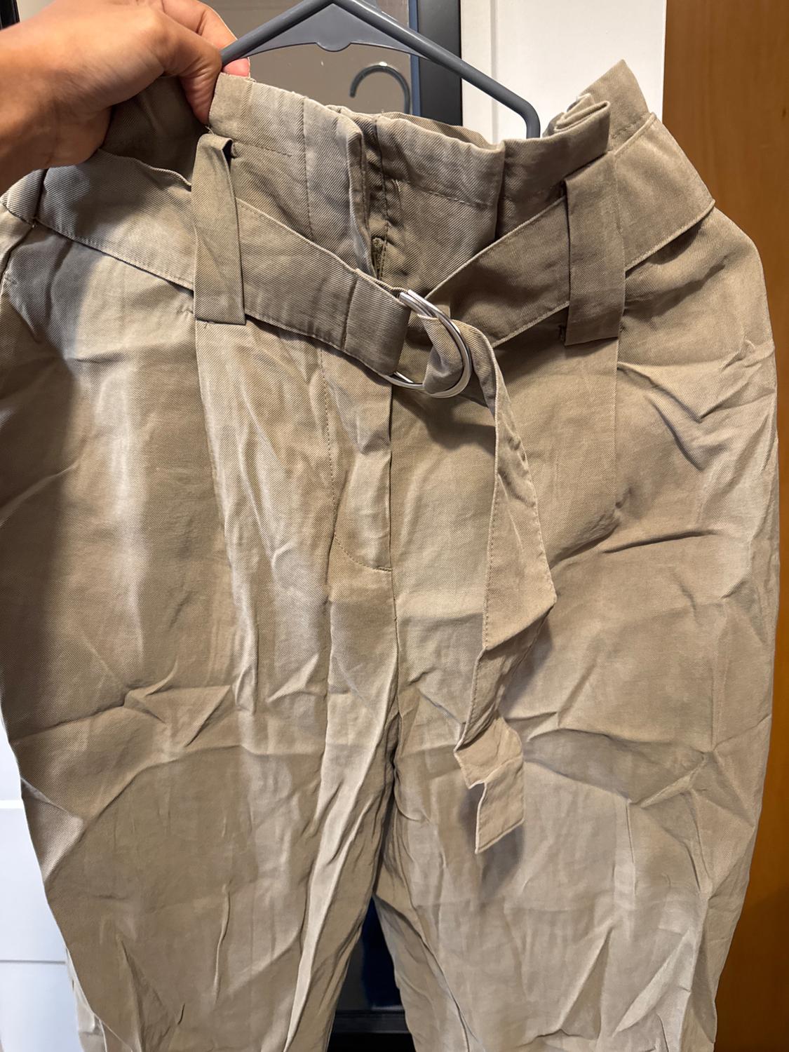 Creased beige trousers hanging on a hanger, with a belt of the same fabric. Interior background of a door and white wall.