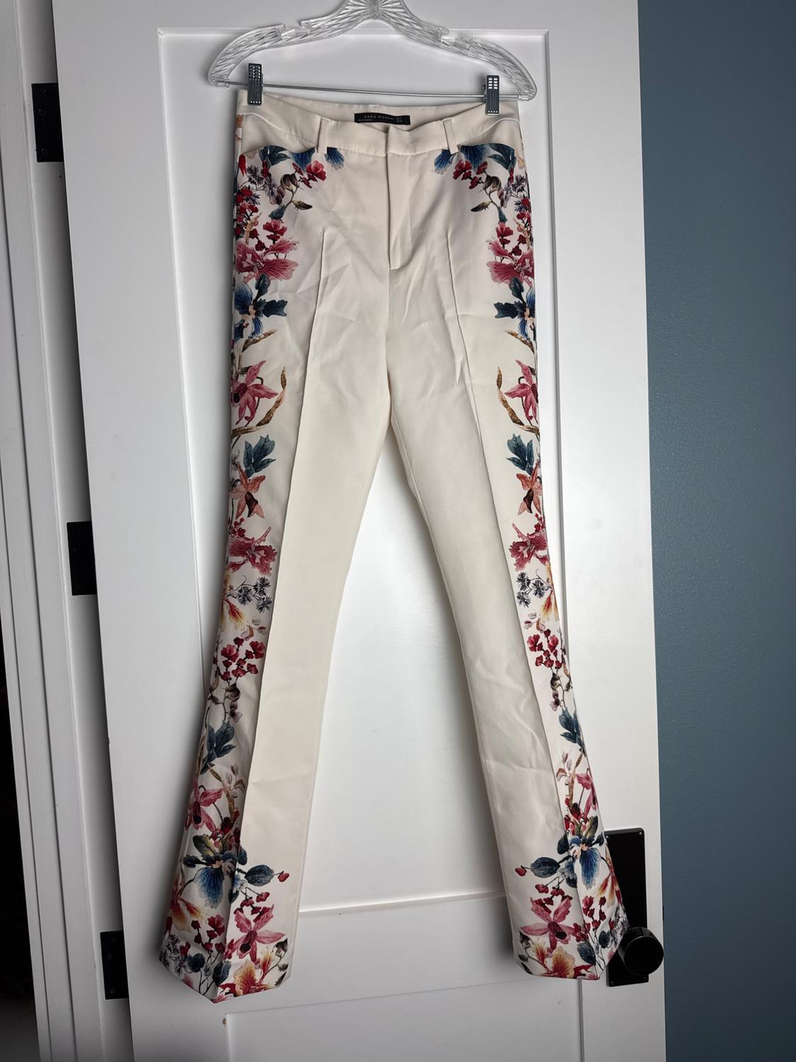 White trousers hanging on a door, featuring a striking red and blue floral print down the sides of both legs.