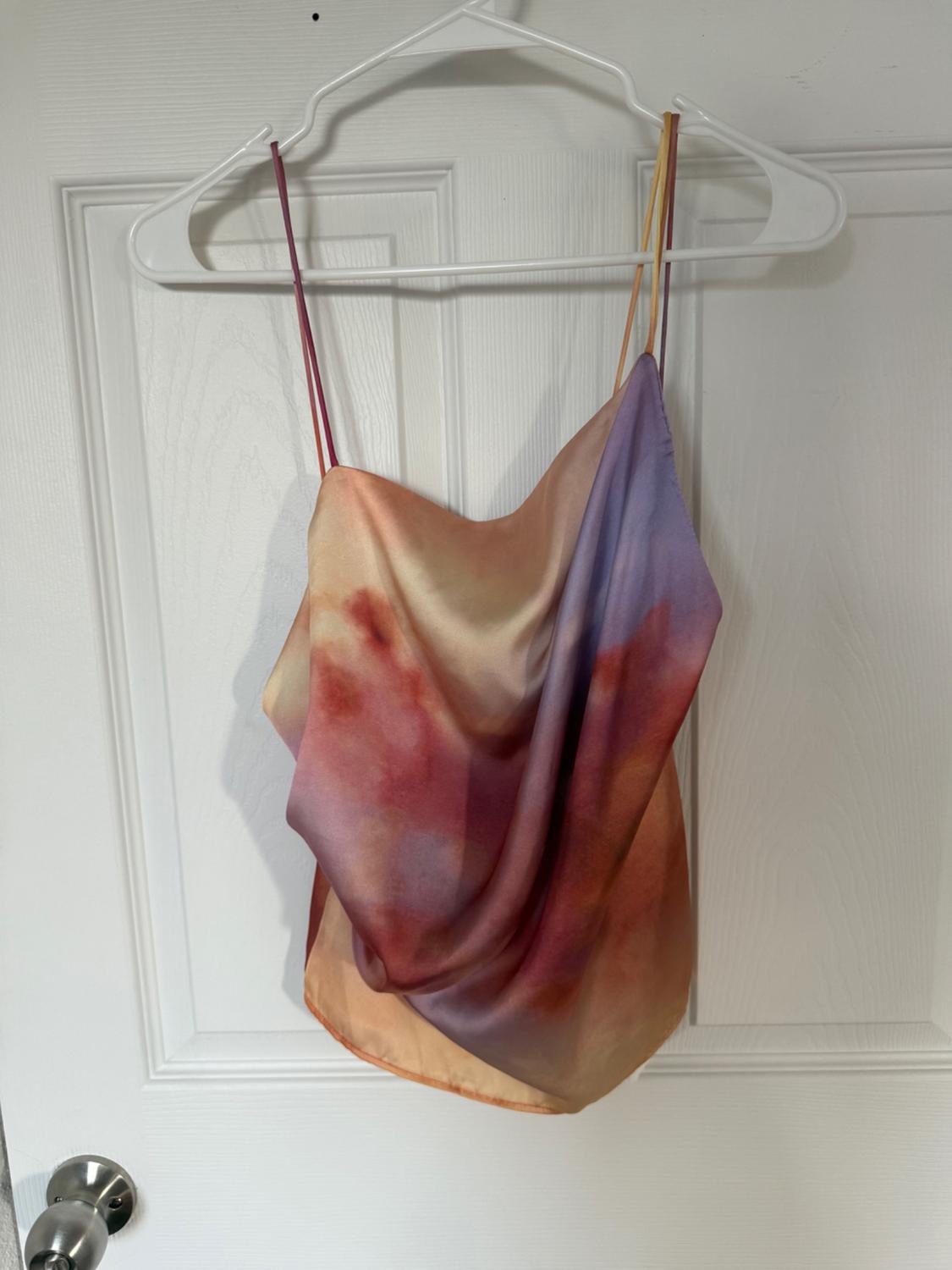 Sleeveless top with thin straps and a gradient print in shades of purple, red and orange, hanging on a white hanger over a white door.