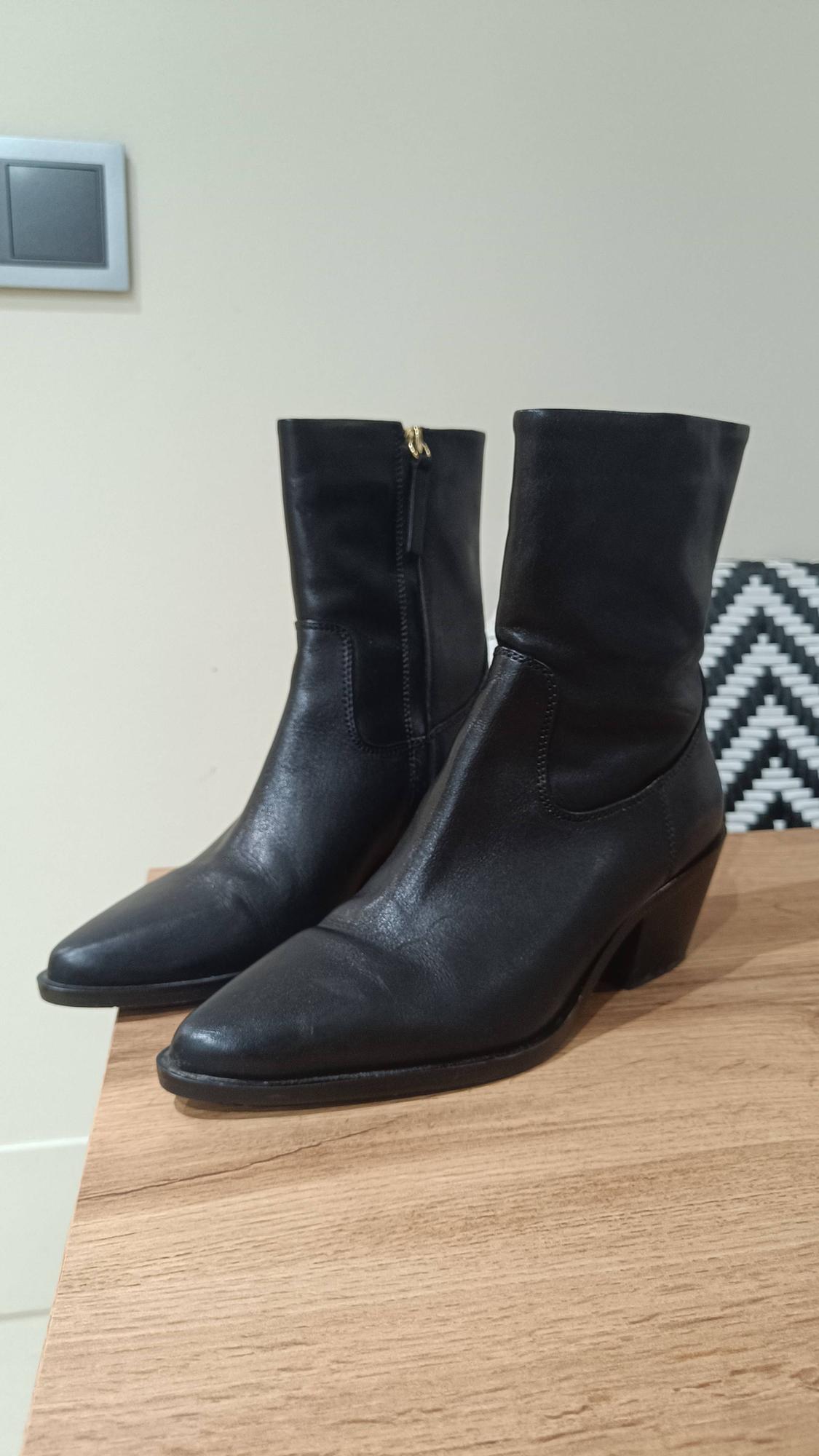 Black leather mid-heel boots on a light wooden table. Background with a beige wall and a geometric pattern cushion.