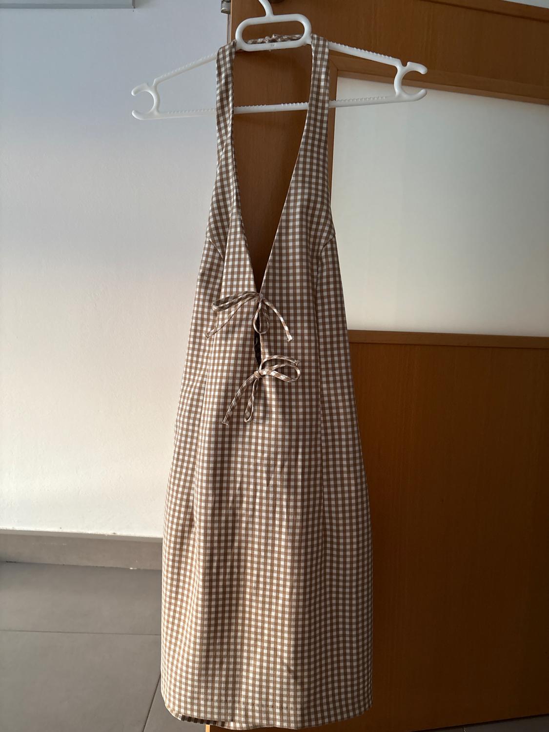 Brown and white checkered dress with straps and a bow on the chest, hanging on a white hanger above a wooden door.
