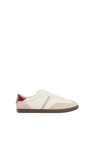 White trainers with beige suede details, a red heel and a brown rubber sole on a white background.