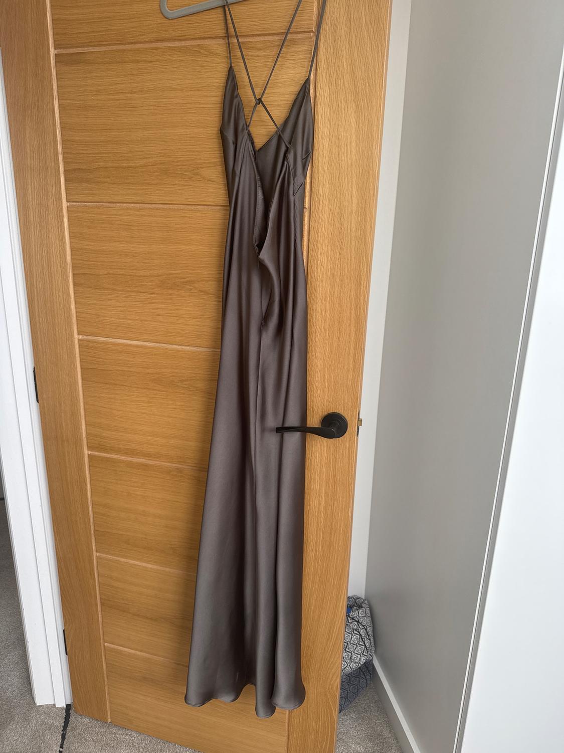 Long grey satin dress hanging on a light wooden door. The design features thin straps crossed at the back.