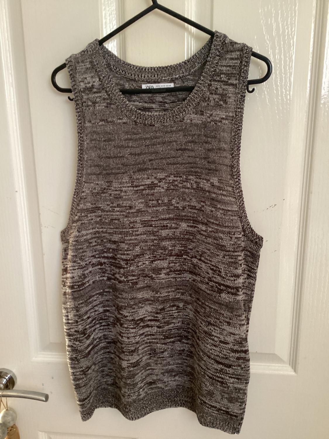 Grey knitted sleeveless top hanging on a black coat hanger over a white door. Label visible on the neck.